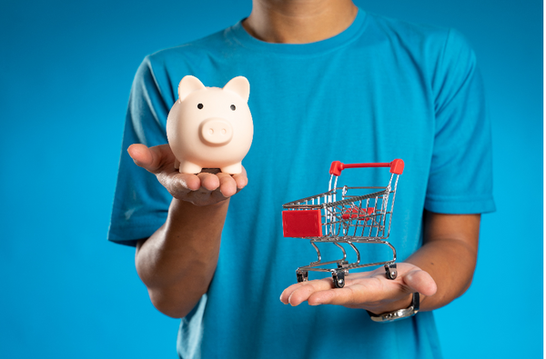 A person in a blue shirt in front of a blue background holding a piggy bank in one hand and a shopping cart in the other 