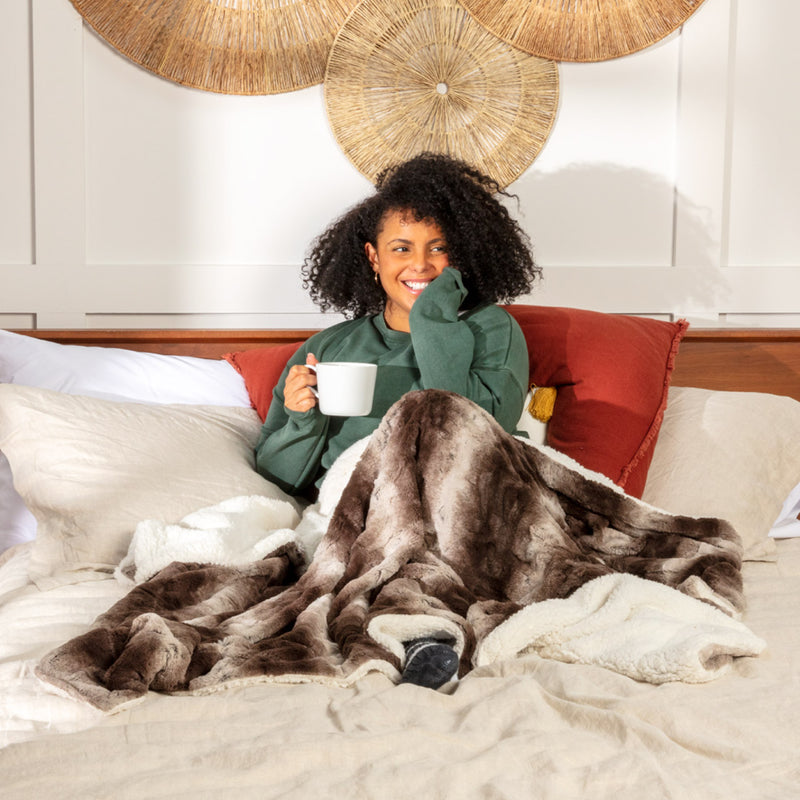 Young woman sitting in bed while wrapped up in a coffee faux fur throw