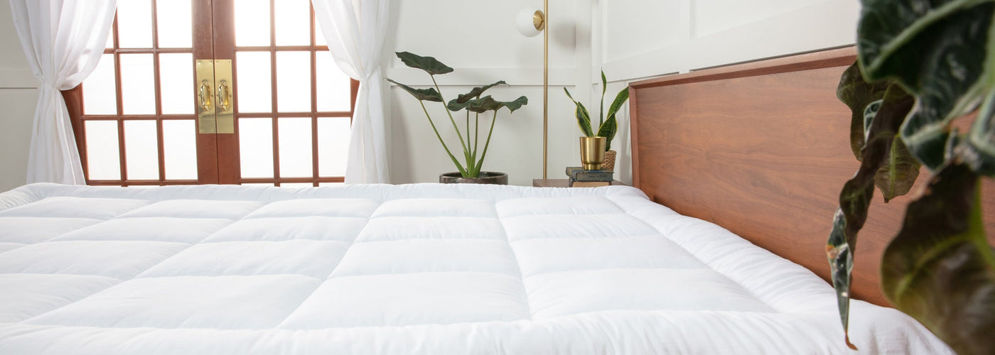 Photo of a white, quilted, pillow top mattress topper on a bed. There is a set of French glass doors with white curtains and a handful of house plants.