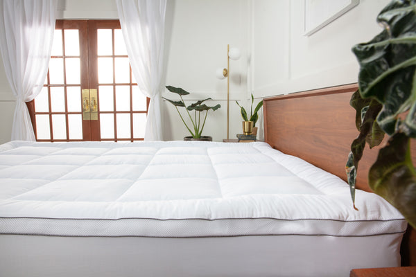Photo of a white, quilted, pillow top mattress topper on a bed. There is a set of French glass doors with white curtains and a handful of house plants.