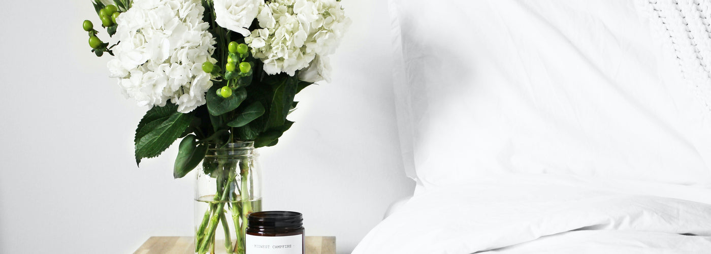 Photo of a vase of white flowers with greenery, plus a candle, on a nightstand next to a bed with white bedding.