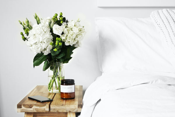Photo of a vase of white flowers with greenery, plus a candle, on a nightstand next to a bed with white bedding.