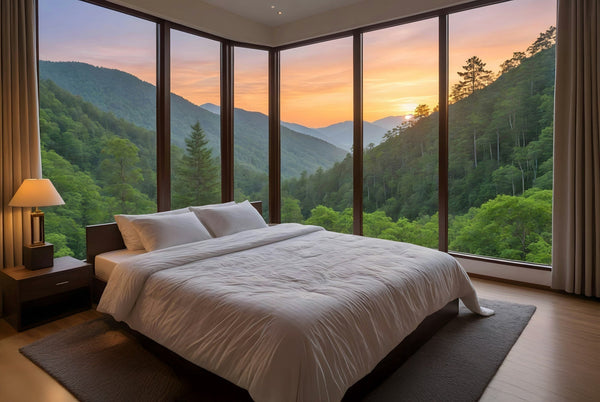 A king size bed with a fluffy white comforter. The bed is positioned in front of floor to ceiling windows, showcasing a beautiful sunset mountain view.