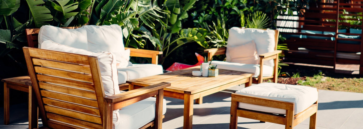 Photo of wooden patio furniture with white cushions surrounded by lush greenery