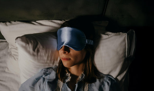 A woman with dark blonde hair sleeping on her back with a light blue satin eye mask over her eyes.