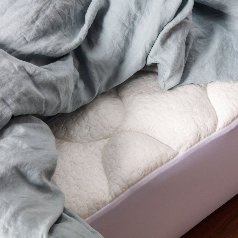 Close up view of a white pillow top mattress pad fitted on a mattress with pale blue bedding on top.